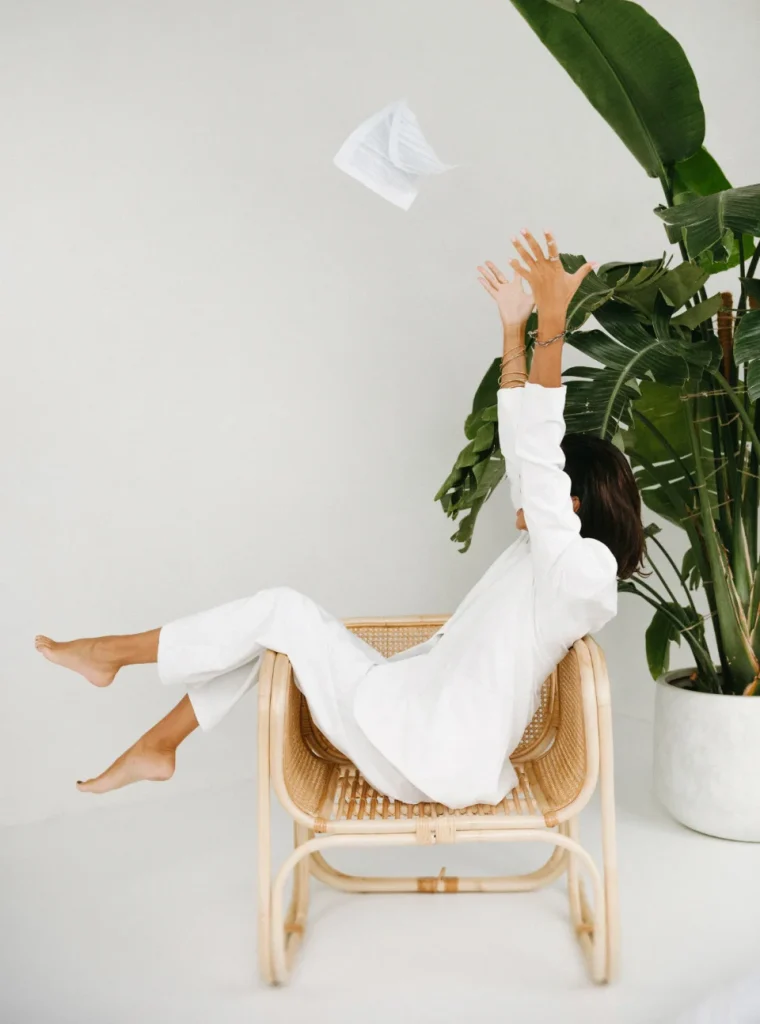 Ewelina Świderska, młoda kobieta w czarnych włosach i mocnym makijażu, siedzi bokiem na ratanowym krześle. biały garnitur. Trzyma obie ręce uniesione do góry, obok zlatują kartki papieru.