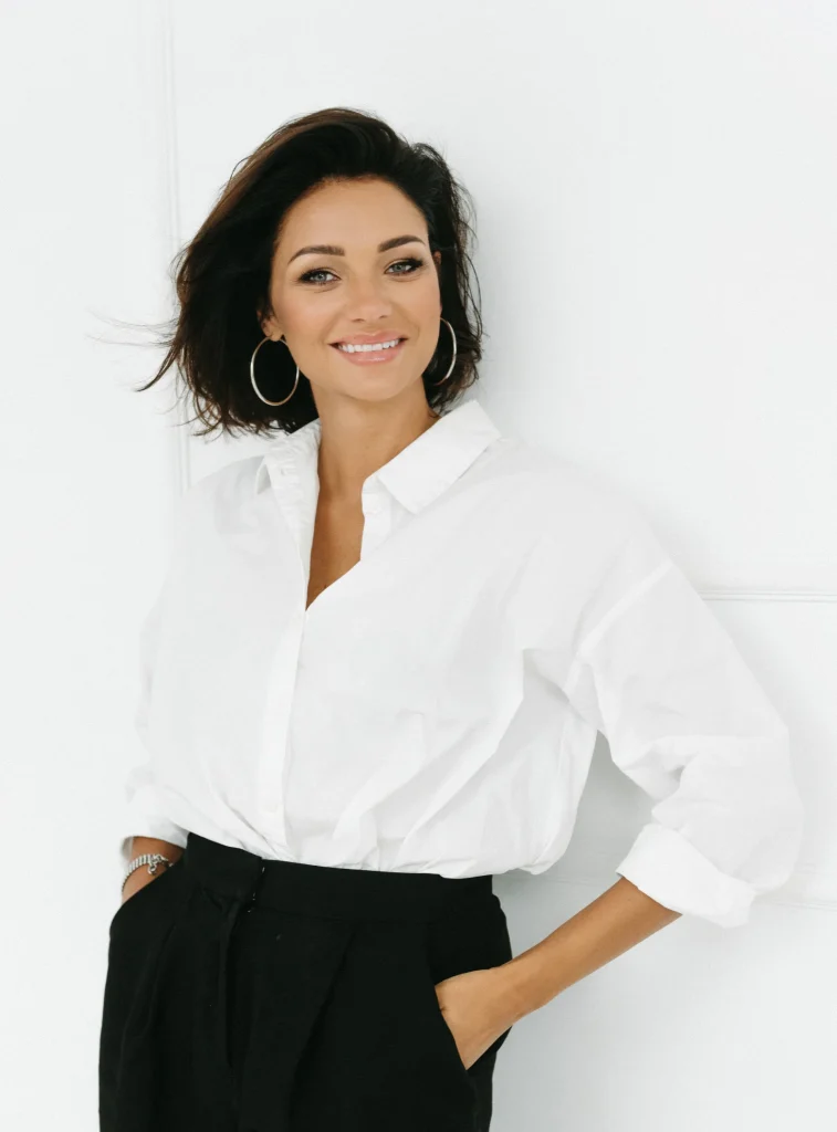 Ewelina Świderska, młoda kobieta w czarnych włosach i mocnym makijażu. Ma na sobie duże kolczyki w kształcie okręgów oraz białą koszulę i czarne eleganckie spodnie. Uśmiecha się szeroko.