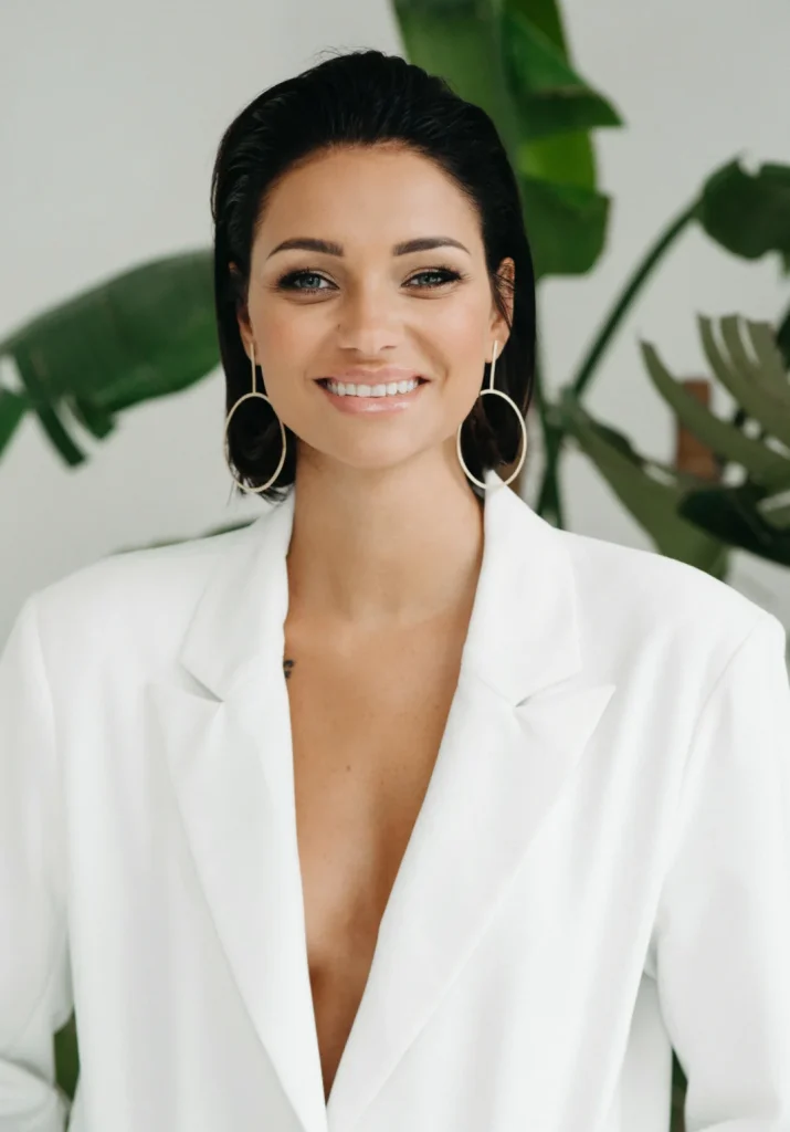 Ewelina Świderska, młoda kobieta w czarnych włosach i mocnym makijażu, stoi na tle zielonej rośliny. Ma na sobie duże kolczyki w kształcie okręgów oraz biały garnitur. Uśmiecha się szeroko.