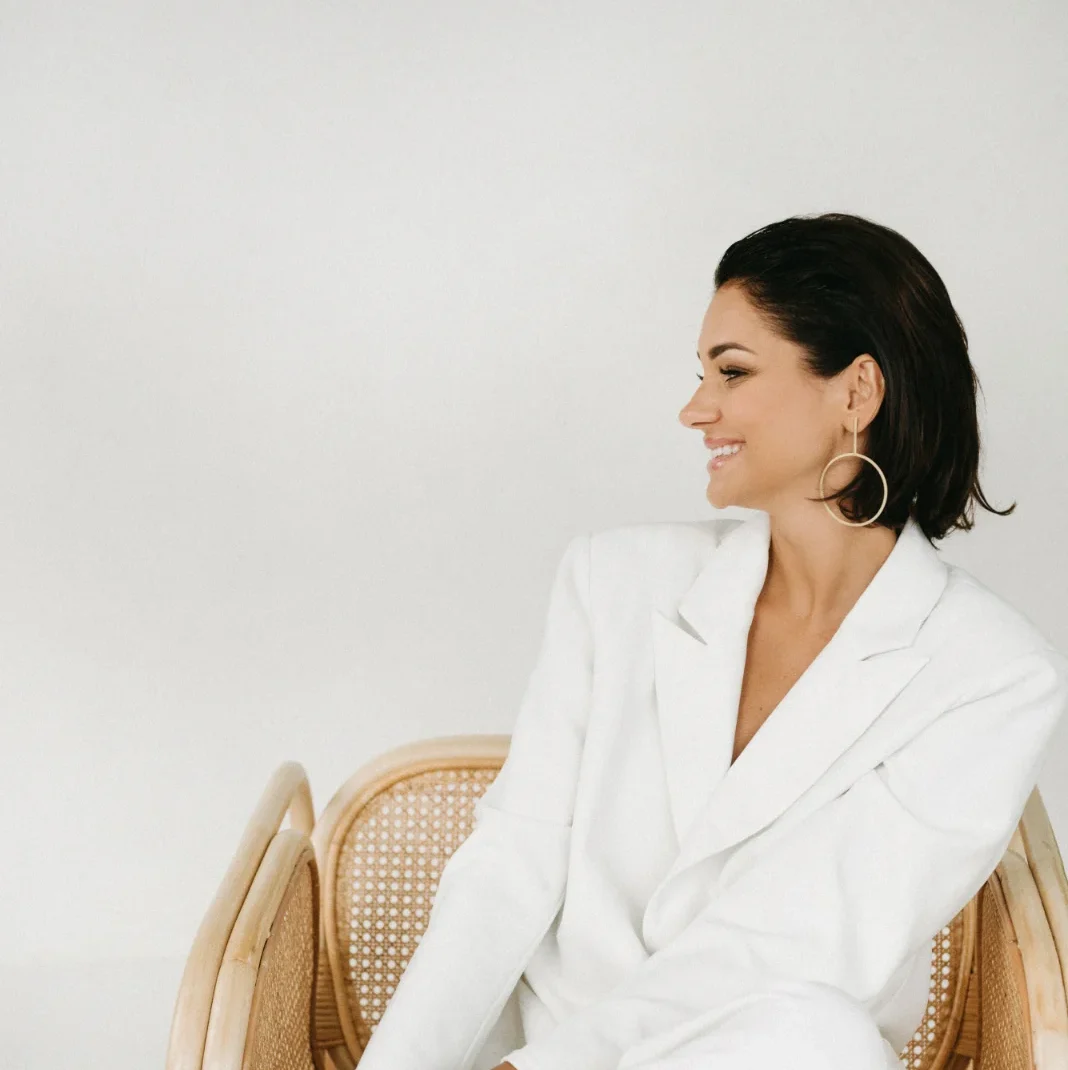 Ewelina Świderska, młoda kobieta w czarnych włosach i mocnym makijażu, siedzi na ratanowym krześle, jest zwrócona w lewą stronę. Ma na sobie duże kolczyki w kształcie okręgów oraz biały garnitur. Uśmiecha się szeroko.