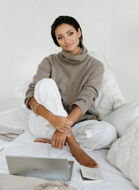 Ewelina Świderska, młoda kobieta w czarnych włosach i mocnym makijażu, siedzi na jasnej kanapie. Ma na sobie duże kolczyki w kształcie okręgów oraz białe szerokie spodnie i beżowy golf. Wokół niej znajduje się otwarty laptop oraz notes.