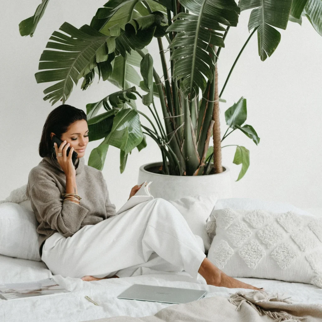 Ewelina Świderska, młoda kobieta w czarnych włosach i mocnym makijażu. Ma na sobie białe szerokie spodnie i beżowy golf. Siedzi na jasnej kanapie, w dłoni trzyma telefon przyłożony do ucha, a w lewej ręce trzyma notes. Ma na sobie dużo pięknej biżuterii, w tle widać dużą zieloną roślinę.